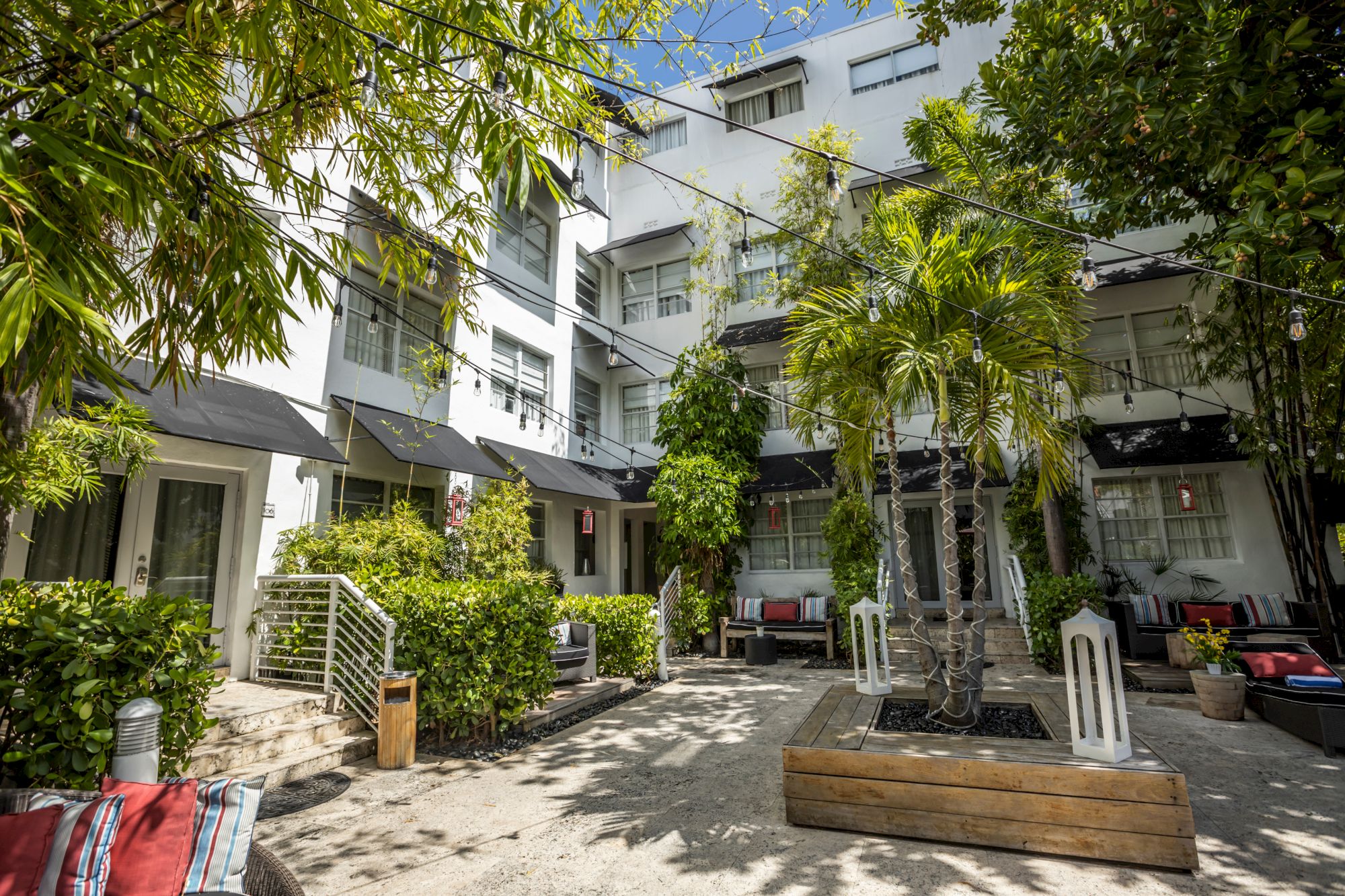 A sunny courtyard of a white multi-story building with balconies, palm trees, shaded seating, planters, and a path through greenery.