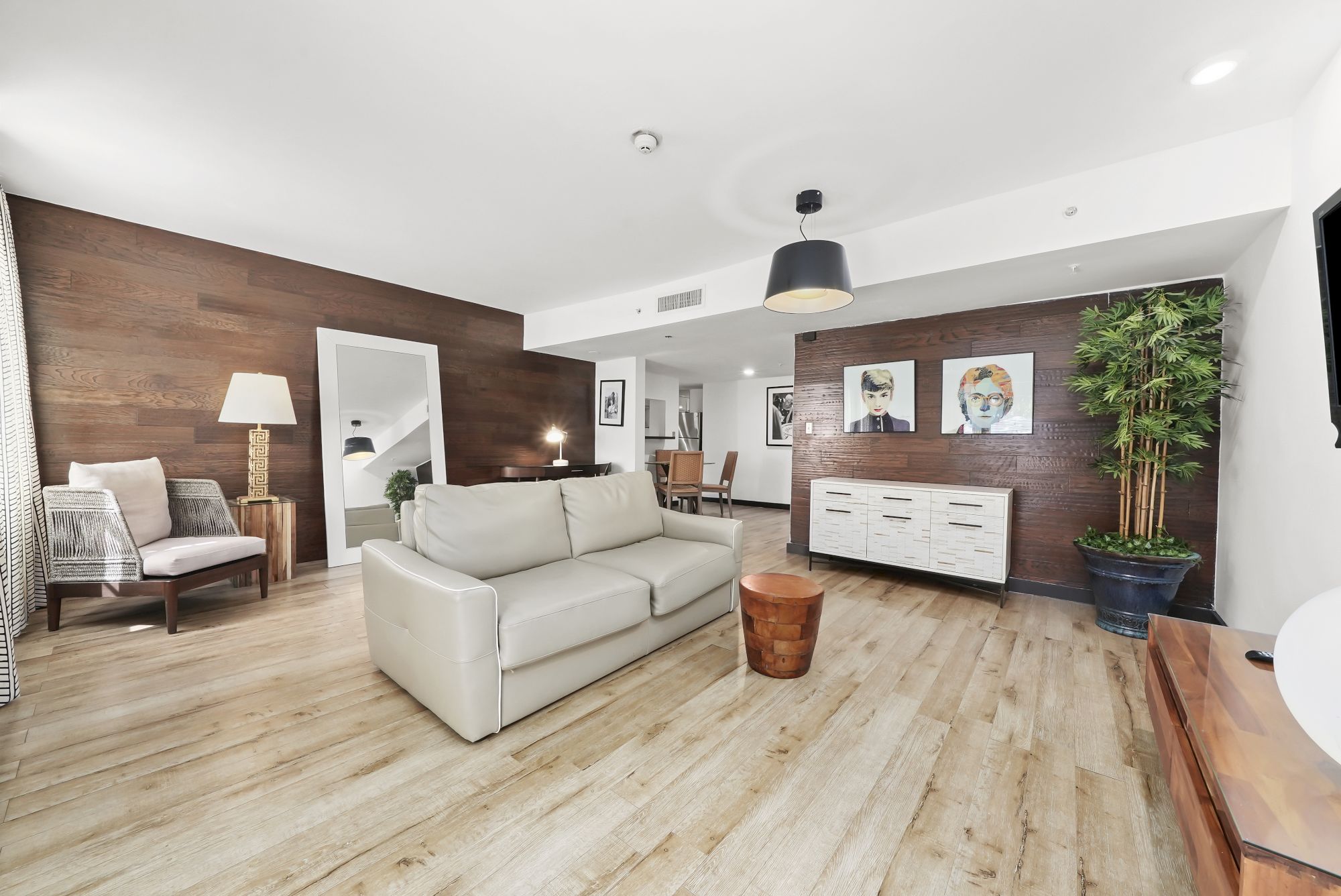 A bright, modern living room with light wood flooring, white sofas, a dark wood accent wall, a white console, potted plant, and artwork.