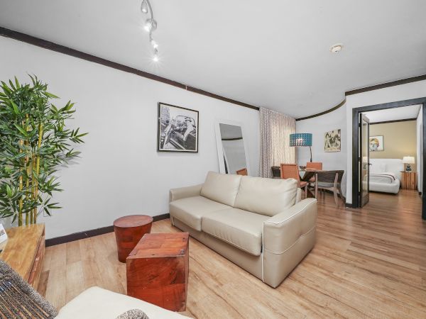 Cozy modern living area with light beige sofa, wooden accents, potted plant, and a desk in the back; open layout leading to a bedroom.