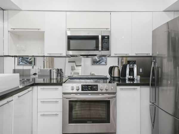 A modern white kitchen with stainless steel appliances, including a fridge, microwave, oven, and coffee station, arranged along glossy white cabinets.