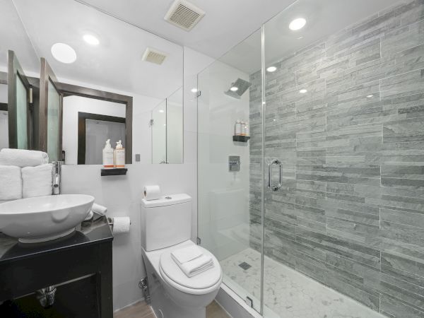 A modern bathroom with a glass-enclosed shower, gray tiled wall, white toilet, pedestal sink, bright lighting, and a dark vanity counter.