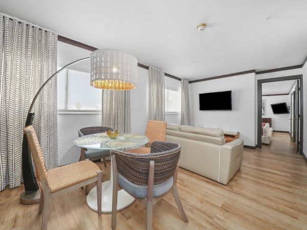 A bright, modern living-dining area with a round glass table, woven chairs, a curved arc floor lamp, beige sofa, wood floor, and a wall-mounted TV.