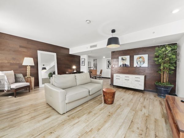 A bright, modern living room with light wood floors, a beige sofa, a dark accent wall, a white sideboard, potted plant, and art on the walls.