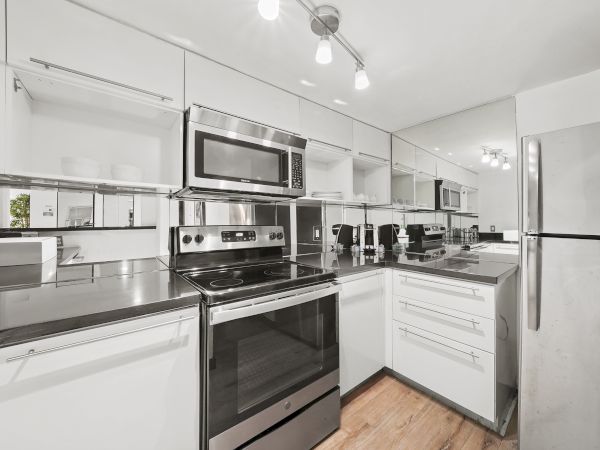 A modern L-shaped kitchen with white cabinets, stainless steel appliances (fridge, microwave, oven), and a dark countertop, well-lit and clean.