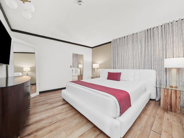 A bright hotel room with a large white bed, pink bed scarf and pillow, a flat-screen TV, wooden floor, and a patterned curtain separating a sitting area.