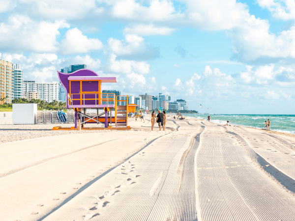 A sunny beach with smooth sand, colorful lifeguard shack, a few people strolling near turquoise waves, and modern buildings lining the coast.