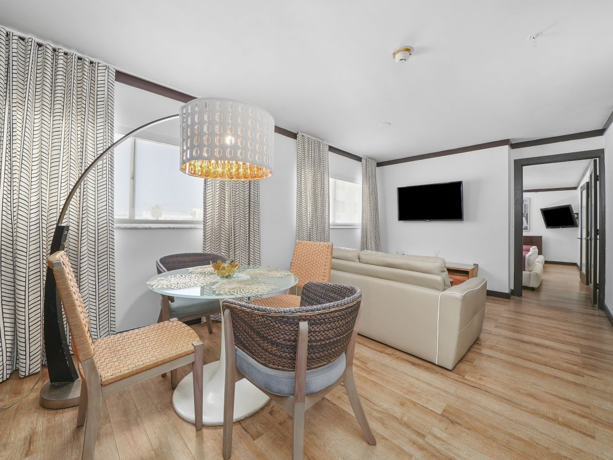 A modern, open-plan living area with a round glass dining table, woven chairs, a curved floor lamp, light wood floors, a beige sofa, and a mounted TV.