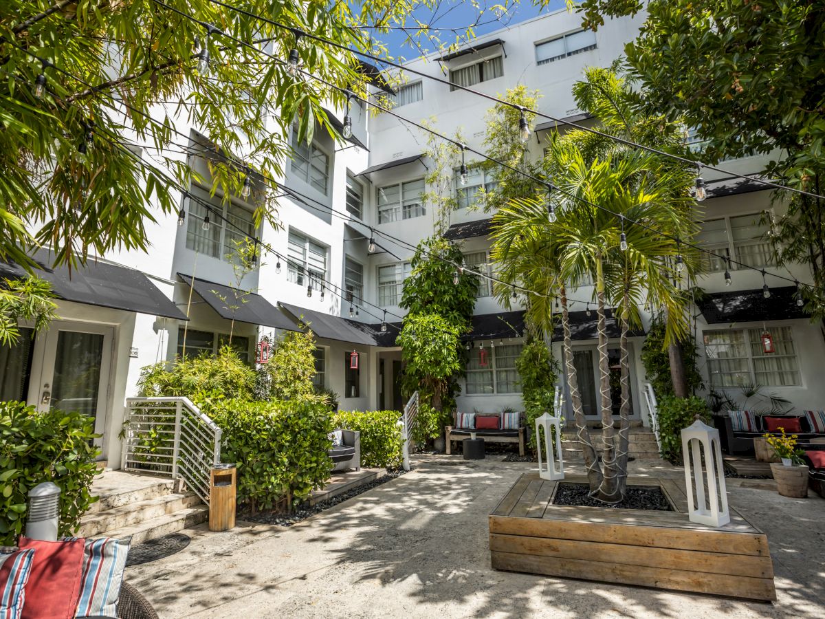 A sunny courtyard surrounded by a three-story residential building, with palm trees, benches, planters, and seating areas.