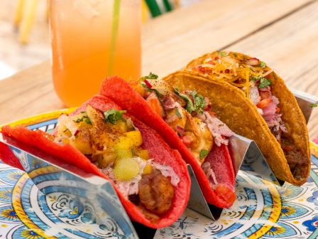 Tacos with grilled meat, shredded cabbage and peppers sit beside a tall orange drink in a jar with a green straw.