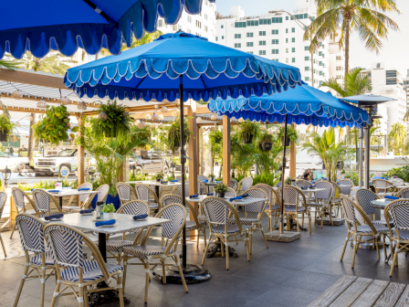 A sunny outdoor dining patio with blue striped awnings, tables and chairs, and palm trees around a hotel courtyard.