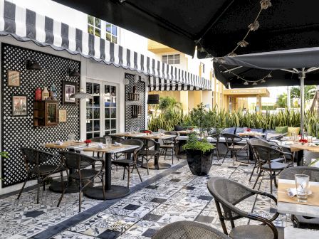 Outdoor restaurant patio with tables, umbrellas, string lights, lush plants, and a sign in the corridor reading &ldquo;Four Freedoms House.&rdquo;