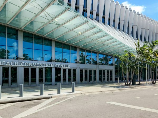 Exterior of the Miami Beach Convention Center with a glass-and-metal canopy, blue sky reflection, and palm trees along the entrance.