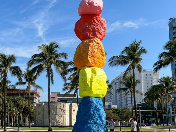 A colorful stacked-stone sculpture stands on a sunny campus with palm trees and pedestrians behind it.