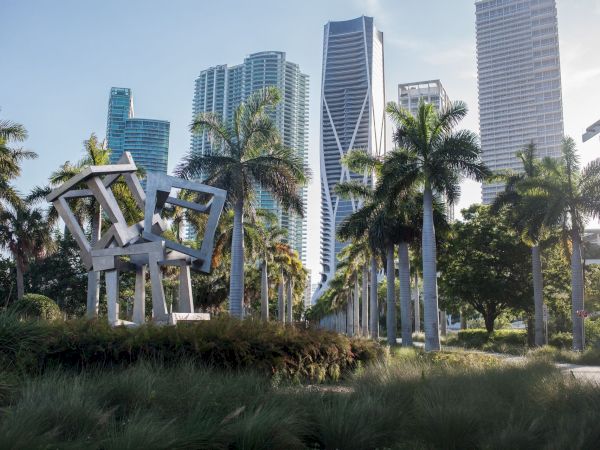 A park with tall palm trees and an abstract metal sculpture, framed by a skyline of modern skyscrapers against a bright sky.