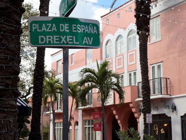 A street sign reads &ldquo;Plaza de Espa&ntilde;a&rdquo; on Drexel Ave, with tall palm trees and string lights outside Spanish-style buildings.