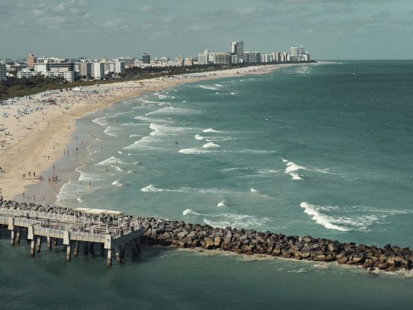 A wide sandy beach with swimmers, waves, a long pier and harbor rocks, plus a distant skyline of tall buildings.