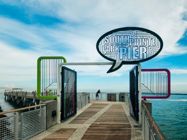 A pier walkway over the water at South Pointe Park, beneath a large &ldquo;South Pointe Park Pier&rdquo; sign, with ocean waves in view.