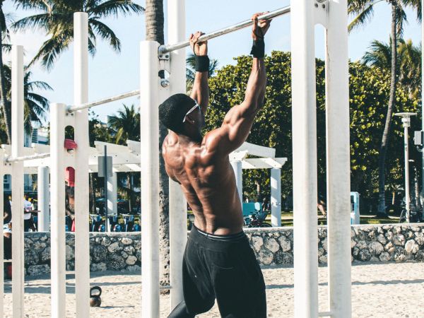 A shirtless man performs pull-ups on a beach workout station, with palm trees and bright sand in the background.