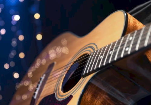A close-up of an acoustic guitar&rsquo;s sound hole and strings with warm bokeh lights in a dark, concert-like background.
