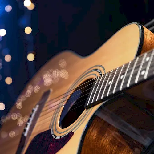 A close-up of an acoustic guitar&rsquo;s sound hole and strings with warm bokeh lights in a dark, concert-like background.