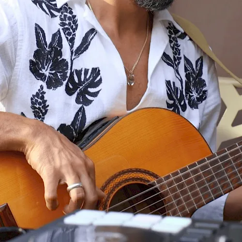 A musician in a floral shirt strums an orange acoustic/classical guitar during a performance.