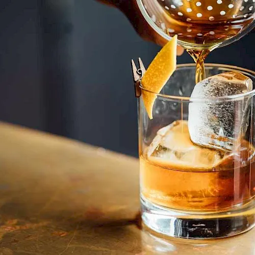 An amber cocktail is being poured over ice in a glass, garnished with a striped orange peel and olive spears.