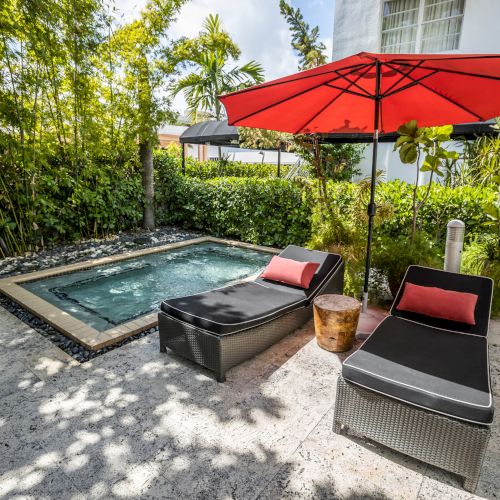 A small backyard pool surrounded by lush greenery, with black lounge chairs and a red patio umbrella shading the patio area.