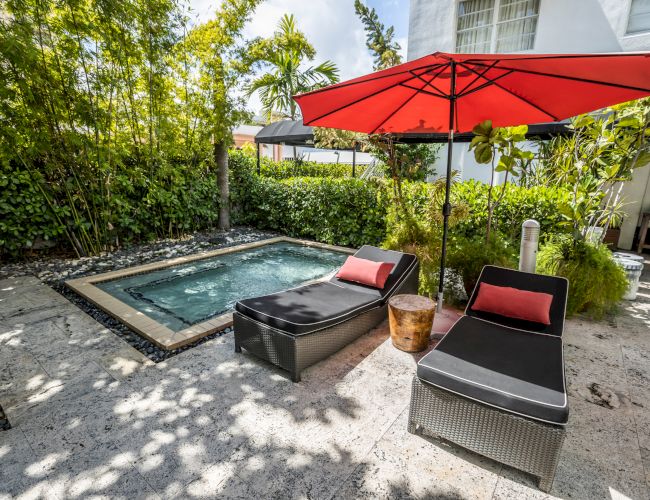 A small backyard pool surrounded by lush greenery, with black lounge chairs and a red patio umbrella shading the patio area.
