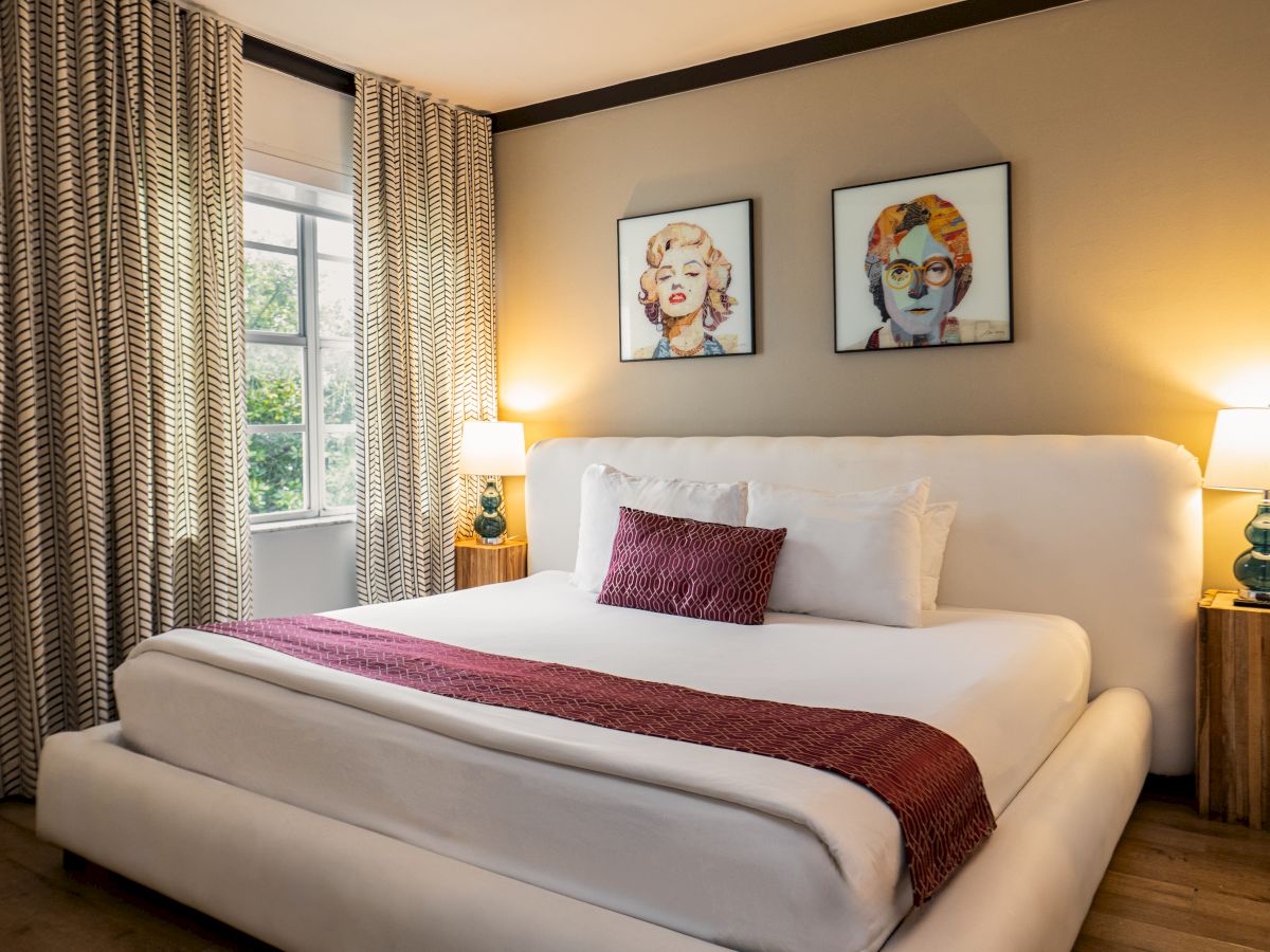 A bright hotel bedroom with a white bed, striped curtains, framed portrait art, and bedside lamps.