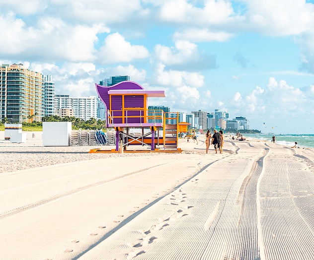 A sunny beach with a colorful lifeguard stand, footprints in sand, people walking, and tall beachfront buildings under blue sky.