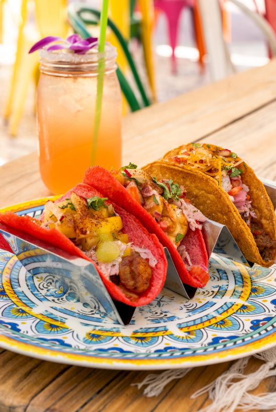 Tacos topped with peppers and herbs sit on a colorful plate beside a jar of orange drink with a green straw.