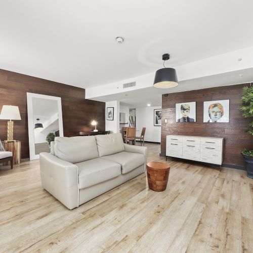 A modern living room with a white sofa, warm wood walls, framed portraits, pendant lights, and indoor plants, all in bright open space.