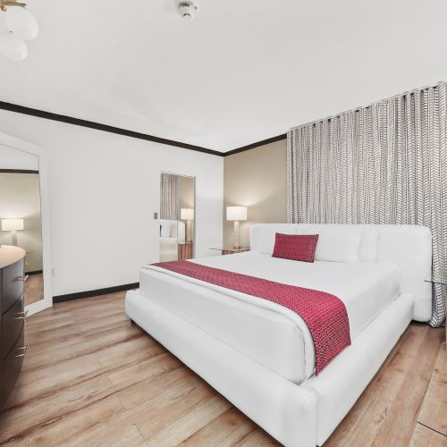 A modern bedroom with a white bed, patterned curtains, bedside lamps, wood floors, and a flat-screen TV.