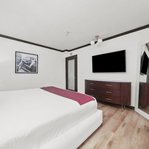 A modern bedroom with a white bed, red runner, wall art, ceiling light, flat-screen TV, dark dresser, and a mirrored wardrobe.