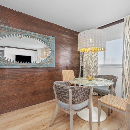 A stylish dining nook with a round table, cushioned wicker chairs, wood-paneled wall, ornate wall mirror, striped curtains, and lamps.